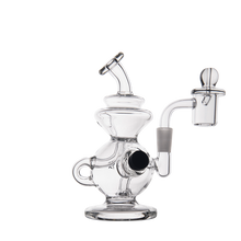 The MJ Arsenal Mini Jig Mini Dab Rig is a clear glass water pipe with a rounded base, side handle, angled mouthpiece, attached bowl, and built-in storage jar. Shown here against a plain white background.
