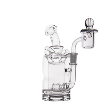 The MJ Arsenal Turbine Mini Dab Rig, made of clear borosilicate glass with a bent neck and cylindrical chamber, features an attached quartz banger and carb cap, shown on a plain white background.