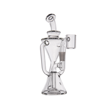 The MJ Arsenal Time Turner Mini Dab Rig is a clear borosilicate glass rig with a uniquely shaped body, multiple angled chambers, a wide stable base, and a cylindrical banger attachment on the right side.