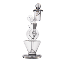 The MJ Arsenal Gemini Mini Dab Rig is a portable clear borosilicate glass apparatus featuring multiple chambers, circular and cylindrical shapes, and a cone-shaped base, shown against a white background.