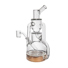 The MJ Arsenal Alpine Series - Apex Mini Rig is a clear glass piece with a cylindrical base, double uptake recycler, multiple chambers, side arm, and angled mouthpiece, displayed on a surface with a wooden accent at the bottom.