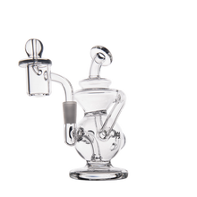 The MJ Arsenal Mini Jig Mini Dab Rig features clear glass, a rounded base, bent neck, side arm recycler design, and a quartz banger on the left. It stands upright against a plain white background.