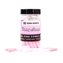 A clear jar labeled “Blazy Susan Pink Paper Cones” with a black lid holds 1 1/4 size pre-rolled pink cones. Several pink cones are displayed in front, and the label notes features like vegan and ultra-thin.