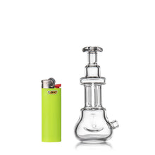 The MJ Arsenal Tetra, a compact, clear borosilicate glass hand pipe, stands upright beside a bright green Bic lighter on a white background, showcasing its modern and portable design.
