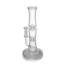 The MJ Arsenal Spire Glass Water Pipe is a clear borosilicate pipe with a wide base, cylindrical neck, multiple chambers, and a 14mm flower bowl, all elegantly shown against a white background.