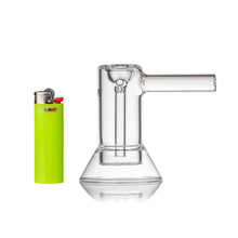 The MJ Arsenal Cove Glass Bubbler, made of transparent borosilicate glass with a cylindrical chamber, stands beside a bright green Bic lighter on a white background.