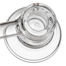 Close-up, top-down view of a clear glass cup with a handle on a round saucer against a white background, evoking the clarity and light reflections of an MJ Arsenal Grotto Glass Pipe crafted from borosilicate glass.