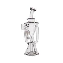 The MJ Arsenal Time Turner Mini Dab Rig is a clear borosilicate glass piece with a conical base, curved tubes, and cylindrical neck with a looped handle, resembling an abstract modern vessel against a white background.