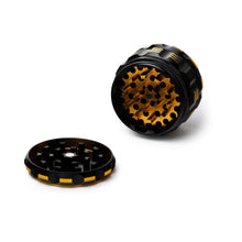 The Human Grade Grinder 5T (2.5") is shown in black and gold with its top and bottom sections separated to reveal sharp aluminum teeth, displayed on a white background.