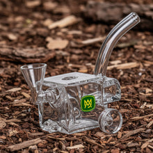 The MJ Arsenal Woodchipper Blunt Bubbler is a clear glass pipe shaped like a small cart, with water filtration for smoother hits, a curved mouthpiece, funnel bowl, two round wheels, and a green and yellow logo on the side.