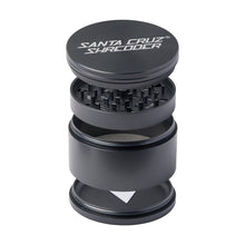 The Santa Cruz Shredder Large 4-Piece Grinder is shown in black anodized aluminum, displayed in an exploded view that reveals its sharp grinding teeth, multiple compartments, and brand name printed on the medical-grade top lid.