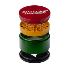 An exploded view of the Santa Cruz Shredder Large 4-Piece Grinder shows its red, yellow, green, and black anodized aluminum parts stacked vertically, highlighting grinding teeth and mesh screen inside.