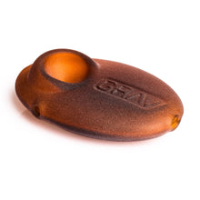 Translucent brown matte Grav Sandblasted Pebble Spoon Pipe with a circular indentation on top and "GRAV" embossed, resembling a sleek art piece, is shown on a white background.