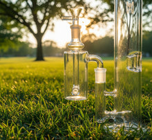 A 14mm DK Barrel Ash Catcher (45° or 90°) is attached to a clear glass bong, resting on green grass in a sunlit park with trees in the background.