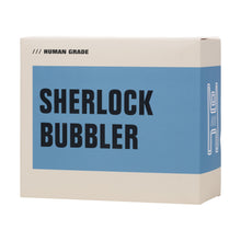 A beige box with a blue label shows bold black text reading Human Grade Sherlock Bubbler, smaller HUMAN GRADE in the top left, and a simple white line drawing of a handmade borosilicate glass piece on the right.