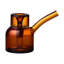The NWTN Home Vesper Bubbler teapot features a modern, minimalist design in translucent amber borosilicate glass, with a cylindrical body, short neck, and long angled spout, elegantly presented on a plain white background.