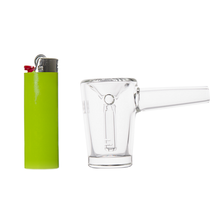 A lime green lighter stands upright next to the MJ Arsenal Basin Bubbler, a clear borosilicate glass pipe with a short stem, both set on a white background.