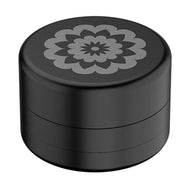 The Flower Mill 2" Next Gen Standard Grinder is a black, cylindrical aluminum grinder with a matte finish and a geometric floral pattern engraved on its lid.