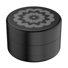 The Flower Mill 2.5" Next Gen Standard Grinder, made of aircraft-grade aluminum in black, features a symmetrical flower-pattern engraving on its flat lid and is displayed against a white background.