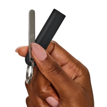 A hand with neatly manicured nails holds the PAX Flow Vaporizer with Grip Sleeve Bundle, featuring a slim vaporizer and a black cylindrical sleeve, against a white background.
