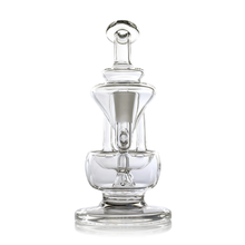 The MJ Arsenal Claude Mini Dab Rig is a clear, intricately designed glass piece featuring a round base, cylindrical body, and looped handle on top—resembling both a decorative dab rig and an elegant art piece.
