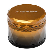 The Human Grade Grinder 7F (2.5") is a metallic, faceted 4-piece aluminum herb grinder with a screw-on lid labeled HUMAN GRADE and features a geometric diamond-like gold and black design for added style.