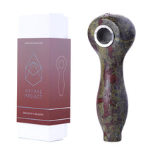 A dark green and red Gemstone Spoon Pipe stands upright beside a white box with a maroon sleeve labeled Astral Project and Dragons Blood, featuring geometric designs and an outline of the pipe.
