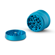 The Human Grade Grinder 3A (2" 4-Piece) is shown open on a white surface, displaying its blue aluminum parts and circular grinding teeth for efficient, reliable herb preparation.