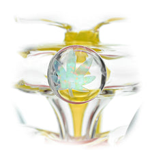 Close-up of the Calibear Loopsicle Recycler Dab Rig, a borosilicate glass piece featuring a round decorative element with an iridescent cannabis leaf inside, set against a translucent yellow-tinted background.