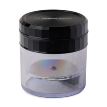 A clear plastic container with a black lid labeled "HUMAN GRADE" holds the Human Grade Storage Grinder 1A (2.5")—a metallic, circular, 4-piece grinder with a shiny iridescent finish inside.