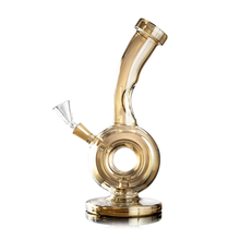 The MJ Arsenal Saturn Water Pipe is a compact, golden-tinted borosilicate glass mini rig with a circular chamber, angled neck, wide base, and left-side clear bowl. It appears clean and is shown against a white background.