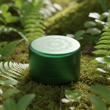 The Flower Mill 2" Next Gen Standard Grinder, featuring a green, round design with a floral lid, rests on moss among ferns and greenery in a natural forest setting.