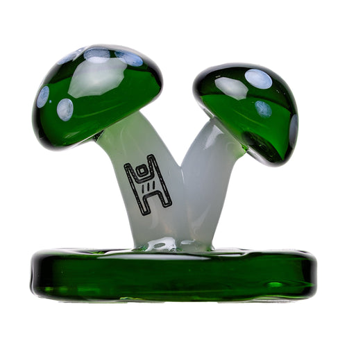 The Mushroom Carb Cap is a borosilicate glass sculpture with two green mushrooms featuring white spots, sharing a translucent stem on a green base. It doubles as a unique carb cap or dab rig accessory and has an abstract symbol on the stem.
