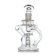 The MJ Arsenal Hydra Mini Dab Rig is a clear glass piece with a unique curvy design inspired by Klein recyclers. It features a grey bowl on top and is shown against a white background.