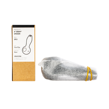 A boxed Grav Classic Spoon Pipe (4-inch, scientific glass) stands next to a gray hand pipe wrapped in white tissue paper and held with a yellow rubber band.