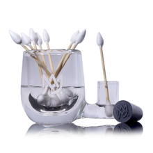 The MJ Arsenal ISO Station, made of clear borosilicate glass with a logo, stores pointed cotton swabs and has an extra swab compartment. A black logo cap sits nearby, all on a reflective surface—perfect for rig cleaning.