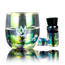 The MJ Arsenal ISO Station is a borosilicate glass smoking pipe with iridescent colors, a rounded bowl, side chamber with black stopper, and white MW logo on the front, shown on a reflective surface.