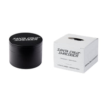 A black Santa Cruz Shredder Large 4-Piece Grinder made of anodized aluminum sits beside its white box labeled "Santa Cruz Shredder" and "Grinder / Broyeur 4 Piece Large," featuring a small cut-out on the top.