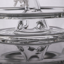 Clear water is poured into a glass dish featuring an artistic centerpiece, evoking the MJ Arsenal Alpine Series - Apex Mini Rig, with swirling reflections and a smooth effect in the transparent container.