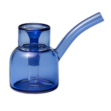 The NWTN Home Vesper Bubbler is a transparent blue borosilicate glass teapot with a wide cylindrical body, matching lid, and long curved spout. Its modern, minimalist design is inspired by the sleek look of the Vesper Bubbler.