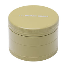 The Human Grade Grinder 4C (2.5") is a round, light olive green aluminum herb grinder with a textured edge, white HUMAN GRADE print on the screw-top lid, and a metal finish for practical use.