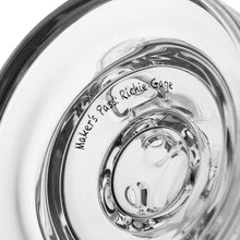 Close-up of the MJ Arsenal The Plasma Core Rig, a clear borosilicate glass piece with smooth, rounded, reflective surfaces and engraved text "Maker’s Pass: Richie Gage" visible.