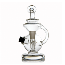 The MJ Arsenal Mini Jig Mini Dab Rig is a clear glass water pipe with a round base, side handle, angled mouthpiece, dome-shaped top, built-in storage jar, and removable black bowl, shown against a white background.