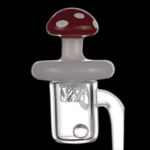 The MJ Arsenal Mushroom Spinner Carb Cap is a glass dab rig attachment with a red and white mushroom-shaped cap, clear cylindrical chamber, and marble detail—an eye-catching accessory for unique dabbing. Shown on a black background.