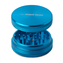 The Human Grade Grinder 1C (2.5" 2-Piece) is a blue aluminum grinder with a textured grip, a partially open lid showing sharp teeth, and "HUMAN GRADE" in silver on top.