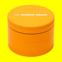 The Human Grade Grinder 4R (2.5") is a bright orange, cylindrical grinder with a screw-on lid, "HUMAN GRADE" in white on top, and a built-in pollen catcher for convenience.