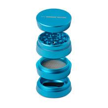 The Human Grade Grinder 3A (2" 4-Piece) in blue aluminum features a lid, grinding chamber, mesh screen, and collection chamber with a scraping tool—perfect for efficient herb preparation.