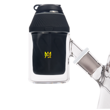 A close-up of an MJ Arsenal Proxy Attachment—a black and yellow piece with a stylized M logo and angled glass joint—crafted from durable borosilicate glass, shown against a minimalistic white background.