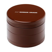 The Human Grade Grinder 4C (2.5") is a brown, round aluminum herb grinder featuring a textured grip and "HUMAN GRADE" printed in white on the lid.