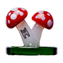 The Mushroom Carb Cap is crafted from borosilicate glass, featuring two red and white spotted mushrooms sharing a single white stem atop a green base with a black geometric design—making it an eye-catching dab rig accessory.